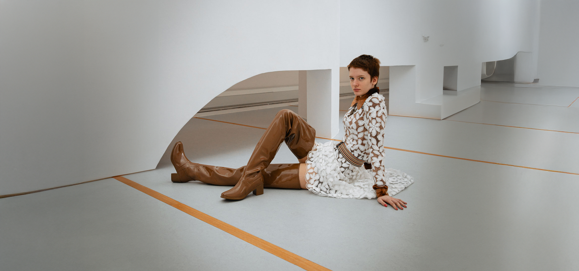 Woman reclining in a minimalist studio wearing Elsa Walnut Brown over-the-knee boots by ACQUO of Sweden styled with a white floral lace dress. Elegant waterproof boots with refined Swedish design, square toes and a sophisticated feminine silhouette in warm walnut brown