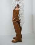 Side view of Elsa Walnut Brown over-the-knee boots by ACQUO of Sweden styled with a white floral lace garment in a minimalist indoor setting. Waterproof boots with elegant Swedish design and sophisticated Scandinavian fashion details.