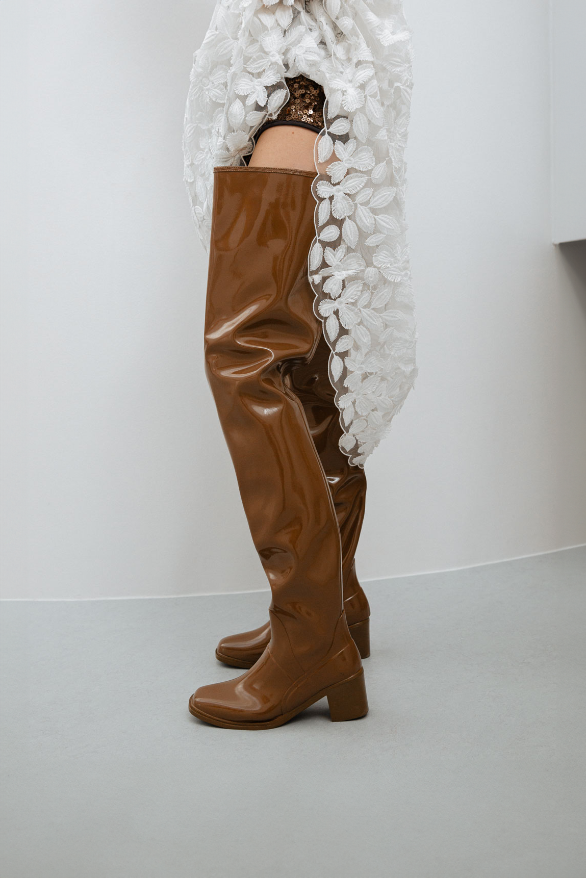 Side view of Elsa Walnut Brown over-the-knee boots by ACQUO of Sweden styled with a white floral lace garment in a minimalist indoor setting. Waterproof boots with elegant Swedish design and sophisticated Scandinavian fashion details.