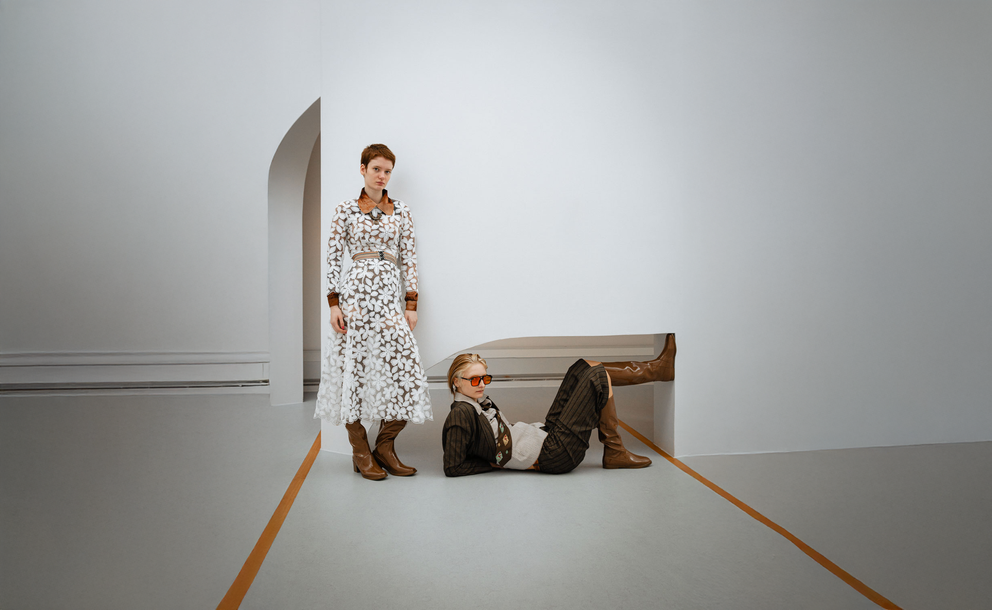 Two women wearing brown knee-high and over-the-knee boots by ACQUO of Sweden in a minimalist architectural interior. Contemporary waterproof boots with square toes and clean Swedish design styled with tailored fashion and a lace dress