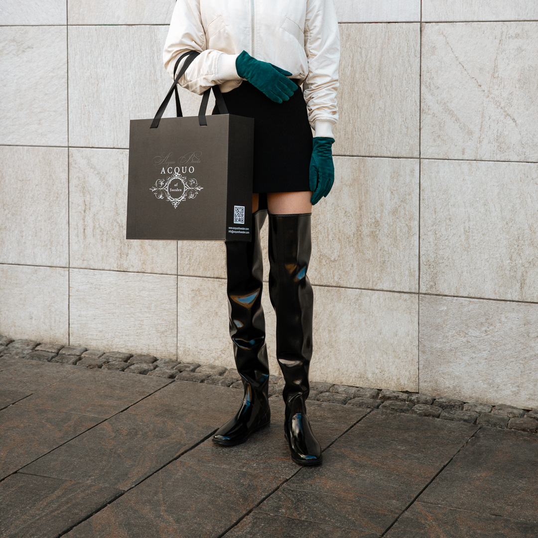 Model wearing ACQUO of Sweden over-the-knee glowing black boots, styled with a cream bomber jacket, black mini skirt, teal gloves, and holding a branded ACQUO shopping bag – a modern and elegant street style look with luxury fashion flair