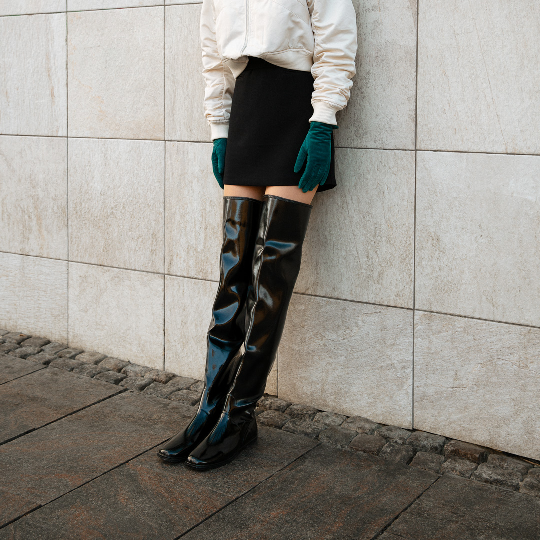 Close-up of ACQUO of Sweden's signature glowing black over-the-knee boots styled with a black mini skirt, ivory bomber jacket, and emerald green gloves, modern Scandinavian fashion combining elegance and attitude in high-shine rubber footwear