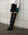 Close-up of ACQUO of Sweden's signature glowing black over-the-knee boots styled with a black mini skirt, ivory bomber jacket, and emerald green gloves, modern Scandinavian fashion combining elegance and attitude in high-shine rubber footwear