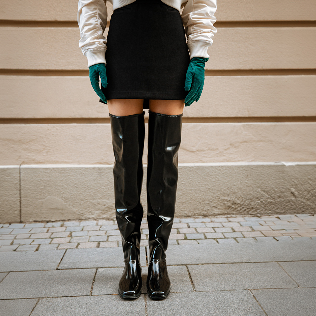 Front view of ACQUO of Sweden’s glowing black over-the-knee rubber boots paired with a black mini skirt, ivory bomber jacket, and emerald green gloves—Scandinavian luxury boots designed for bold, fashion-forward expression