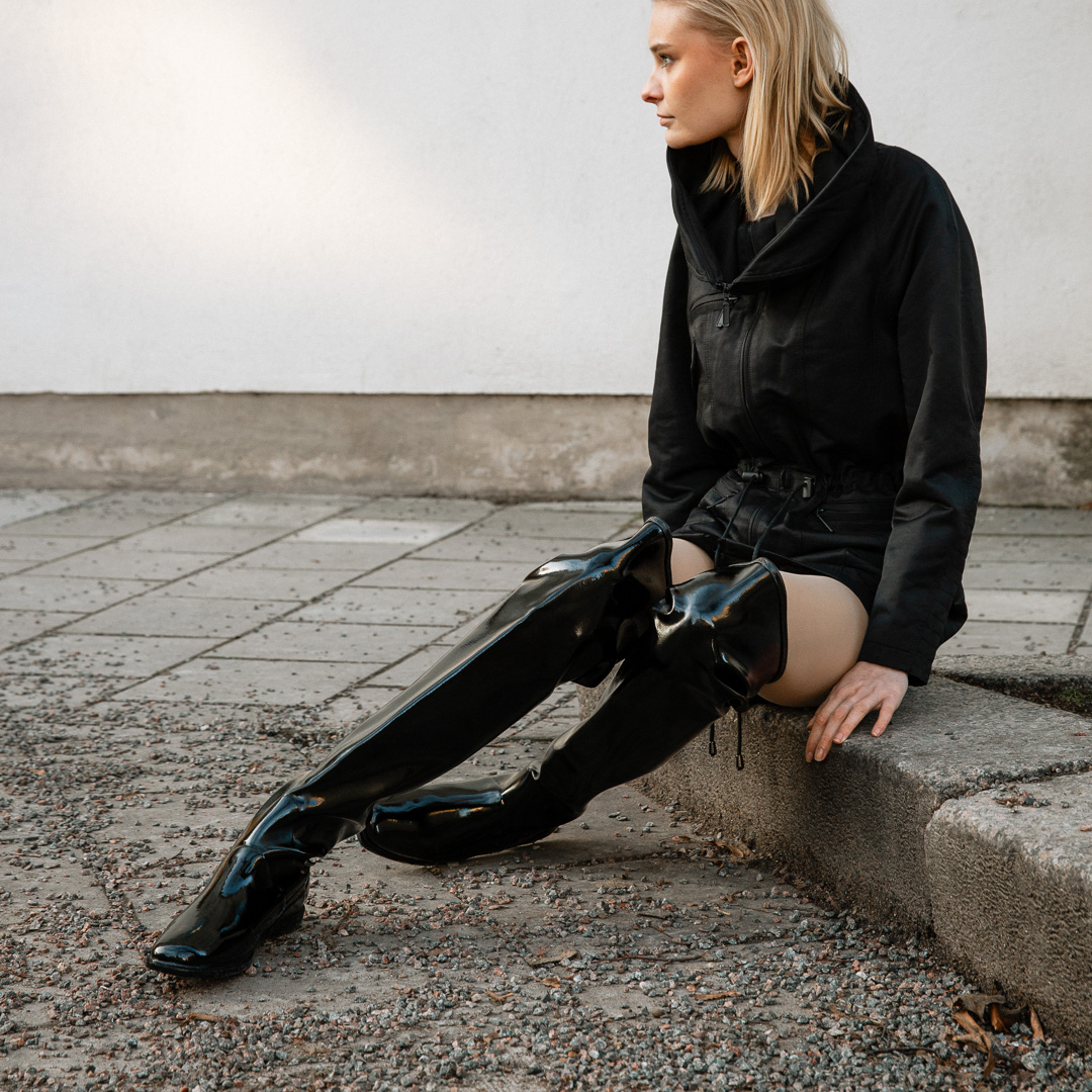 Model in urban street setting wearing ACQUO of Sweden’s black over the knee rubber boots and edgy black zippered jacket, showcasing minimalist Swedish design and fashion-forward footwear perfect for modern style lovers.