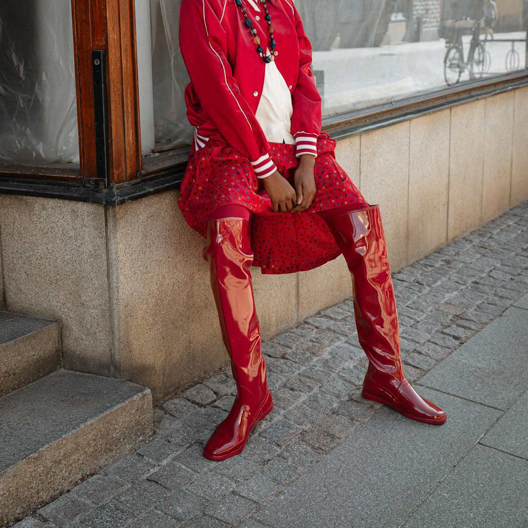 Model wearing Greta Cherry Red over-the-knee boots by ACQUO of Sweden styled with a red skirt and satin bomber jacket in an urban setting. Waterproof boots with elegant Swedish design and modern Scandinavian fashion appeal.