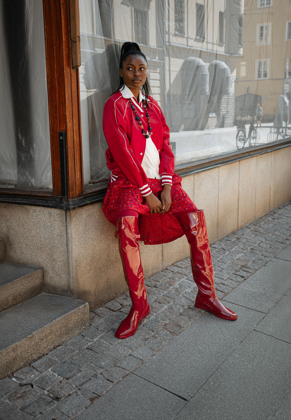 Model wearing Greta Cherry Red over-the-knee boots by ACQUO of Sweden styled with a red satin jacket and patterned skirt on a city street. Waterproof boots featuring elegant Swedish design and contemporary Scandinavian fashion.