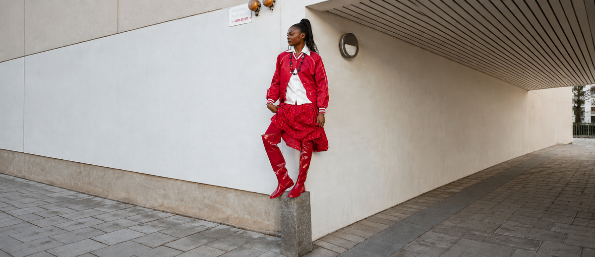 Model posing with one foot raised on a stone post, showcasing ACQUO of Sweden Cherry Red over-the-knee natural rubber boots, styled with a red patterned skirt, white shirt and red jacket against a minimalist urban backdrop