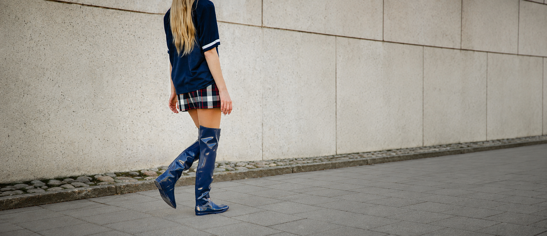Model walking along a city pavement wearing blue over-the-knee natural rubber boots from ACQUO of Sweden, styled with a navy top and short checked skirt for a contemporary Swedish fashion look.