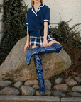 Model sitting on a large stone outdoors wearing Greta Iris Blue boots by ACQUO of Sweden. The image features iris blue over-the-knee rubber boots with a sleek reflective finish, styled with a navy blouse, plaid mini skirt and pearl necklace beside ivy-covered concrete walls and cobblestones.