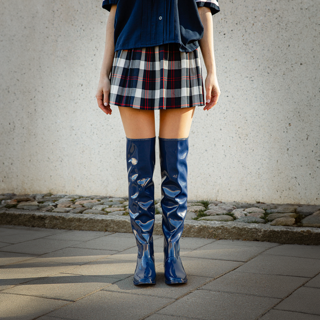 Front view of a model standing outdoors wearing Greta Iris Blue boots by ACQUO of Sweden. The image features iris blue over-the-knee rubber boots with a sleek reflective finish, styled with a navy shirt and plaid mini skirt against a textured concrete wall and cobblestone pavement.