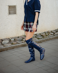 Side view of a model walking outdoors in Greta Iris Blue boots by ACQUO of Sweden. The image shows iris blue over-the-knee rubber boots with a sleek reflective finish, styled with a navy blouse and plaid skirt on a city pavement beside a textured stone wall