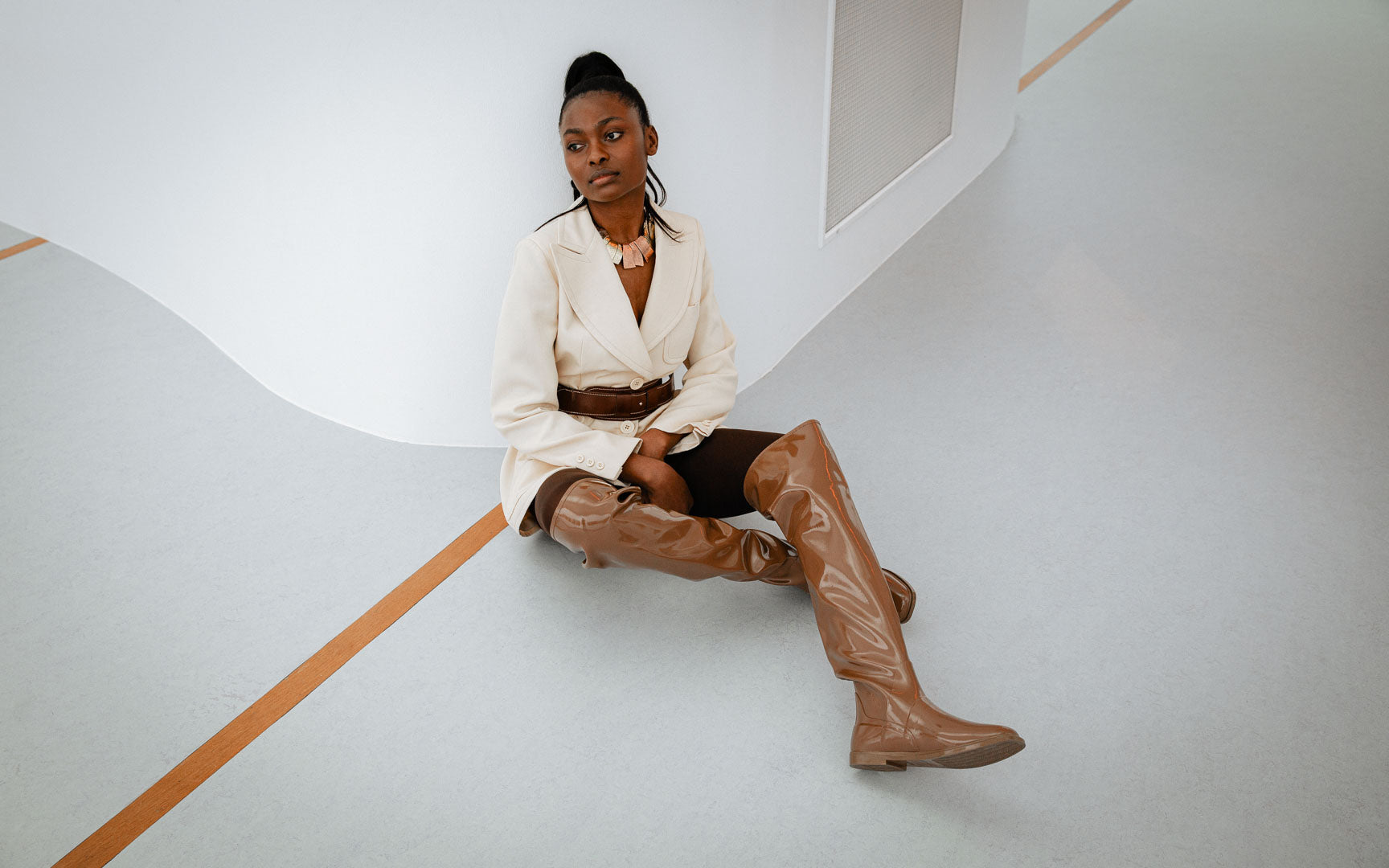 Model seated on the floor with her legs crossed, wearing brown over-the-knee natural rubber boots from ACQUO of Sweden, styled with a cream blazer and brown shorts in a minimalist modern interior.