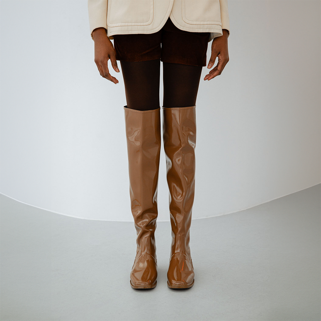 Front view of Greta Walnut Brown knee-high boots by ACQUO of Sweden styled with dark tights, tailored shorts and a cream blazer in a minimalist indoor setting. Elegant waterproof boots with refined Swedish design and a clean contemporary silhouette.