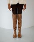 Front view of Greta Walnut Brown knee-high boots by ACQUO of Sweden styled with dark tights, tailored shorts and a cream blazer in a minimalist indoor setting. Elegant waterproof boots with refined Swedish design and a clean contemporary silhouette.