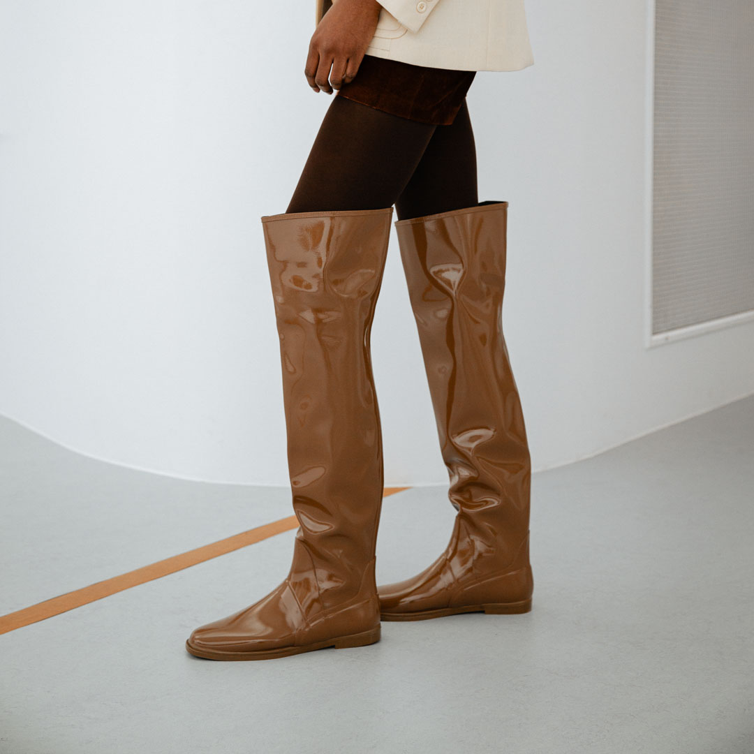 Side view of Greta Walnut Brown knee-high boots by ACQUO of Sweden styled with dark tights and a cream blazer in a minimalist indoor setting. Elegant waterproof boots with refined Swedish design and a sleek modern silhouette.