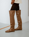 Side view of Greta Walnut Brown knee-high boots by ACQUO of Sweden styled with dark tights and a cream blazer in a minimalist indoor setting. Elegant waterproof boots with refined Swedish design and a sleek modern silhouette.