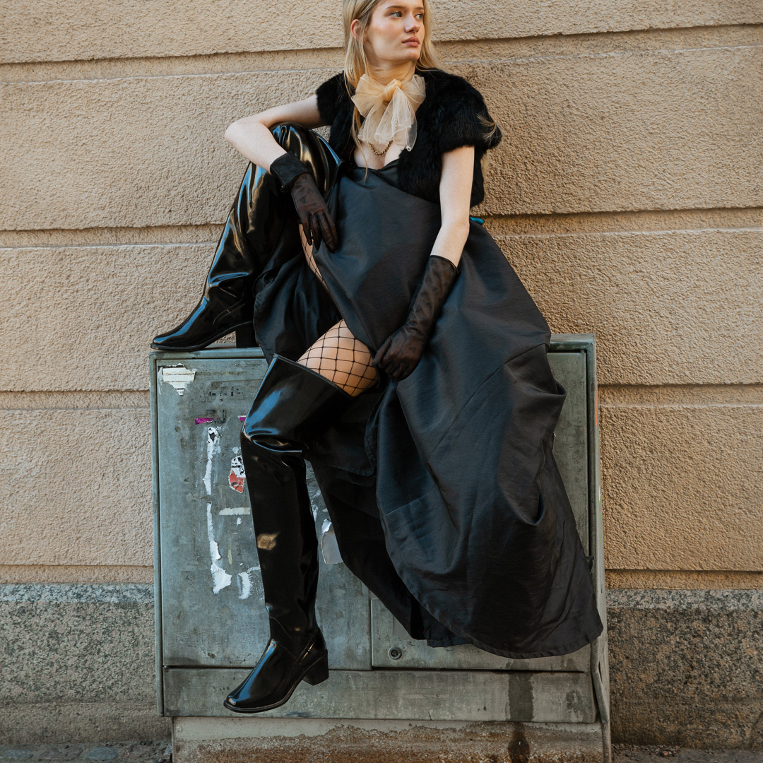 Model wearing ACQUO of Sweden over-the-knee glowing black boots with heels, styled with a dramatic black gown, fishnet stockings, sheer gloves, and a beige tulle neck bow – a bold blend of high fashion and urban edge in a Scandinavian city setting
