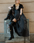 Model wearing ACQUO of Sweden over-the-knee glowing black boots with heels, styled with a dramatic black gown, fishnet stockings, sheer gloves, and a beige tulle neck bow – a bold blend of high fashion and urban edge in a Scandinavian city setting