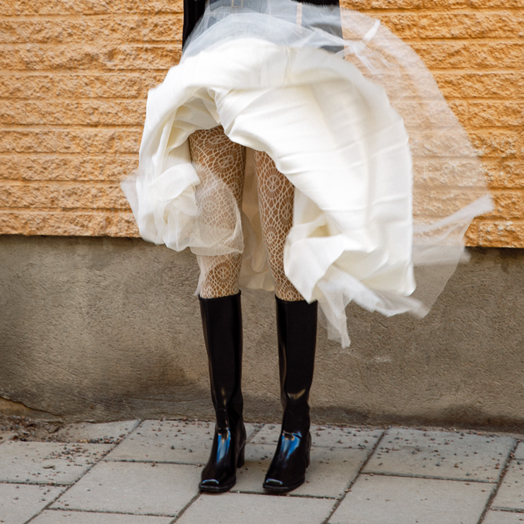 Woman wearing knee-high black rubber boots from ACQUO of Sweden paired with patterned white tights and a flowing layered white skirt, captured in motion against a rustic stone wall—an elegant fusion of vintage and modern Scandinavian style