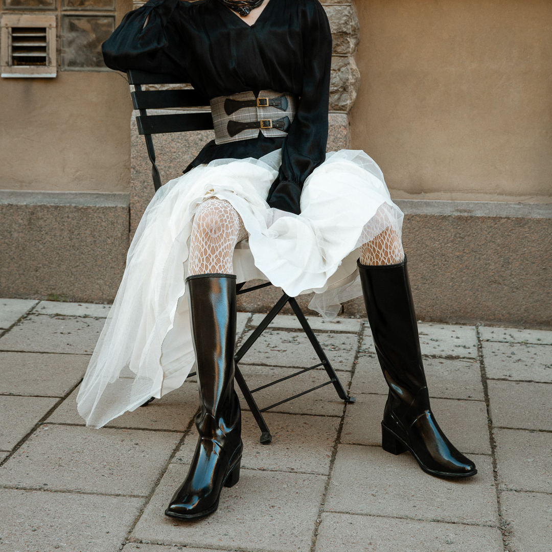 Model in ACQUO of Sweden's knee-high sleek shine black boots with a block heel, paired with a flowing white tulle skirt, textured white tights, and a sleek black blouse – blending feminine elegance with edgy street style