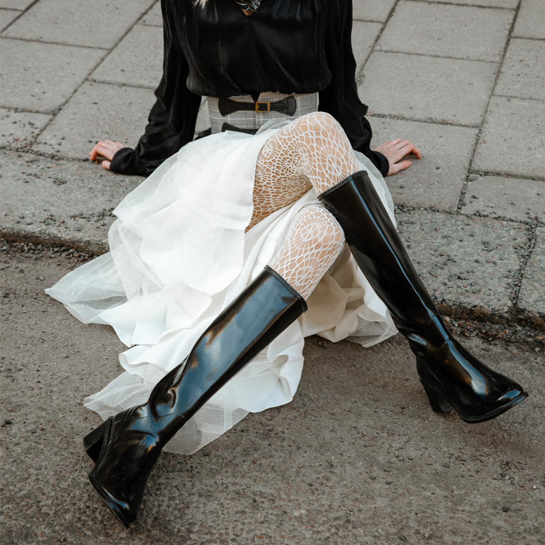 Model wearing ACQUO of Sweden knee-high glowing black boots with block heel, styled with a white layered tulle skirt, patterned white tights, and a black long-sleeve top – a bold and elegant street style look