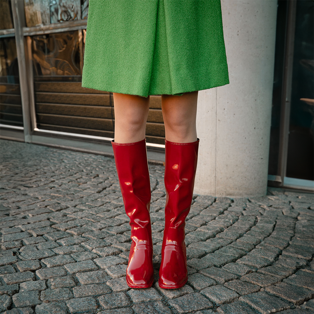 Model wearing Karin Cherry Red knee-high boots by ACQUO of Sweden styled with a green coat on a cobblestone street. Waterproof boots featuring elegant Swedish design and timeless Scandinavian fashion