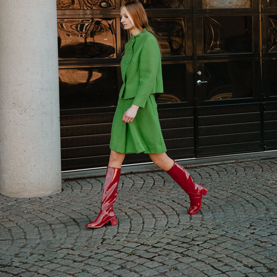 Model wearing Karin Cherry Red knee-high boots by ACQUO of Sweden styled with a green tailored outfit on a cobblestone street. Waterproof boots with elegant Swedish design and refined Scandinavian fashion styling