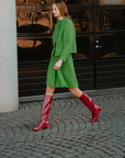 Model wearing Karin Cherry Red knee-high boots by ACQUO of Sweden styled with a green tailored outfit on a cobblestone street. Waterproof boots with elegant Swedish design and refined Scandinavian fashion styling