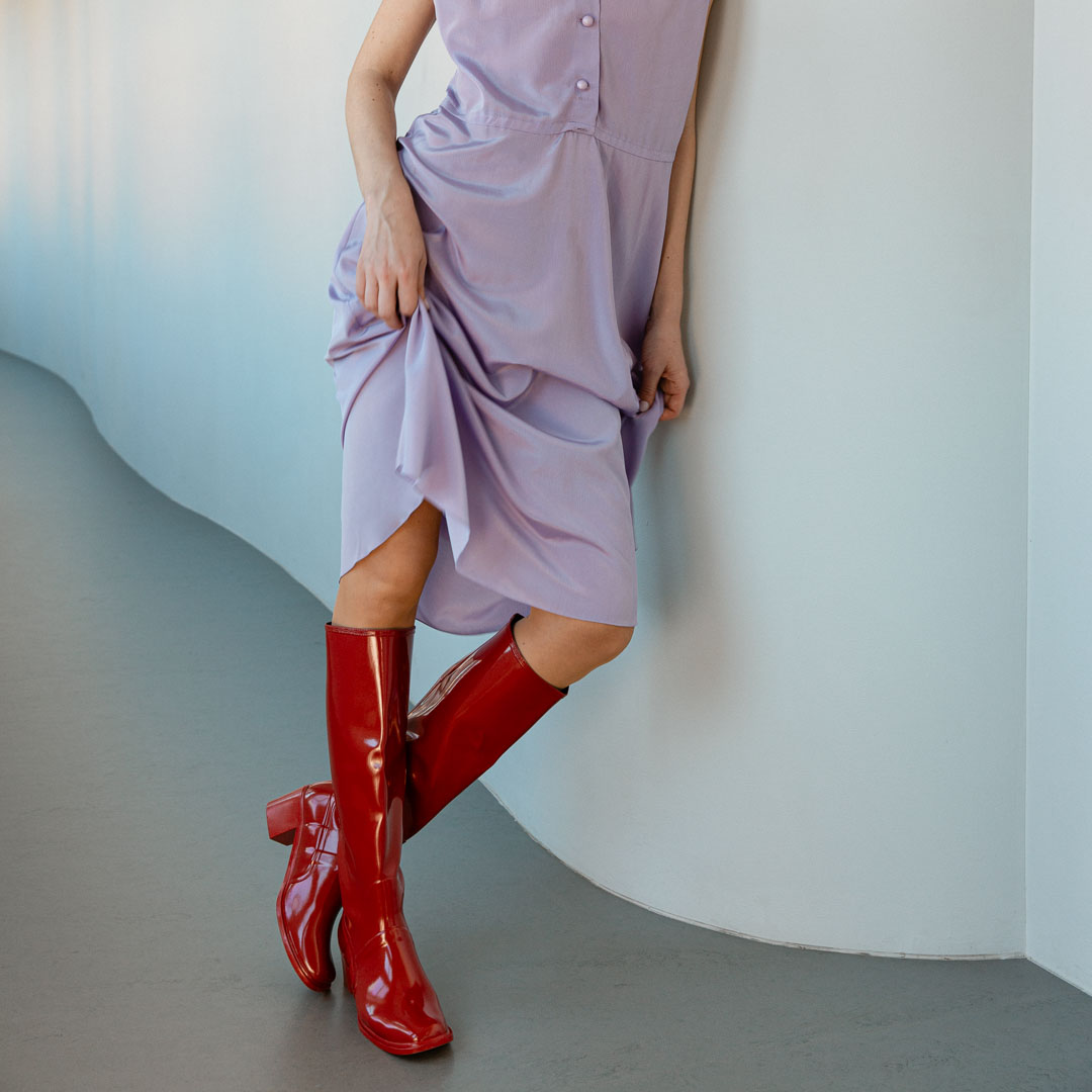 Model wearing Karin Cherry Red knee-high boots by ACQUO of Sweden styled with a soft lavender dress in a modern interior. Waterproof boots featuring elegant Swedish design and minimalist Scandinavian fashion.