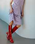 Model wearing Karin Cherry Red knee-high boots by ACQUO of Sweden styled with a soft lavender dress in a modern interior. Waterproof boots featuring elegant Swedish design and minimalist Scandinavian fashion.