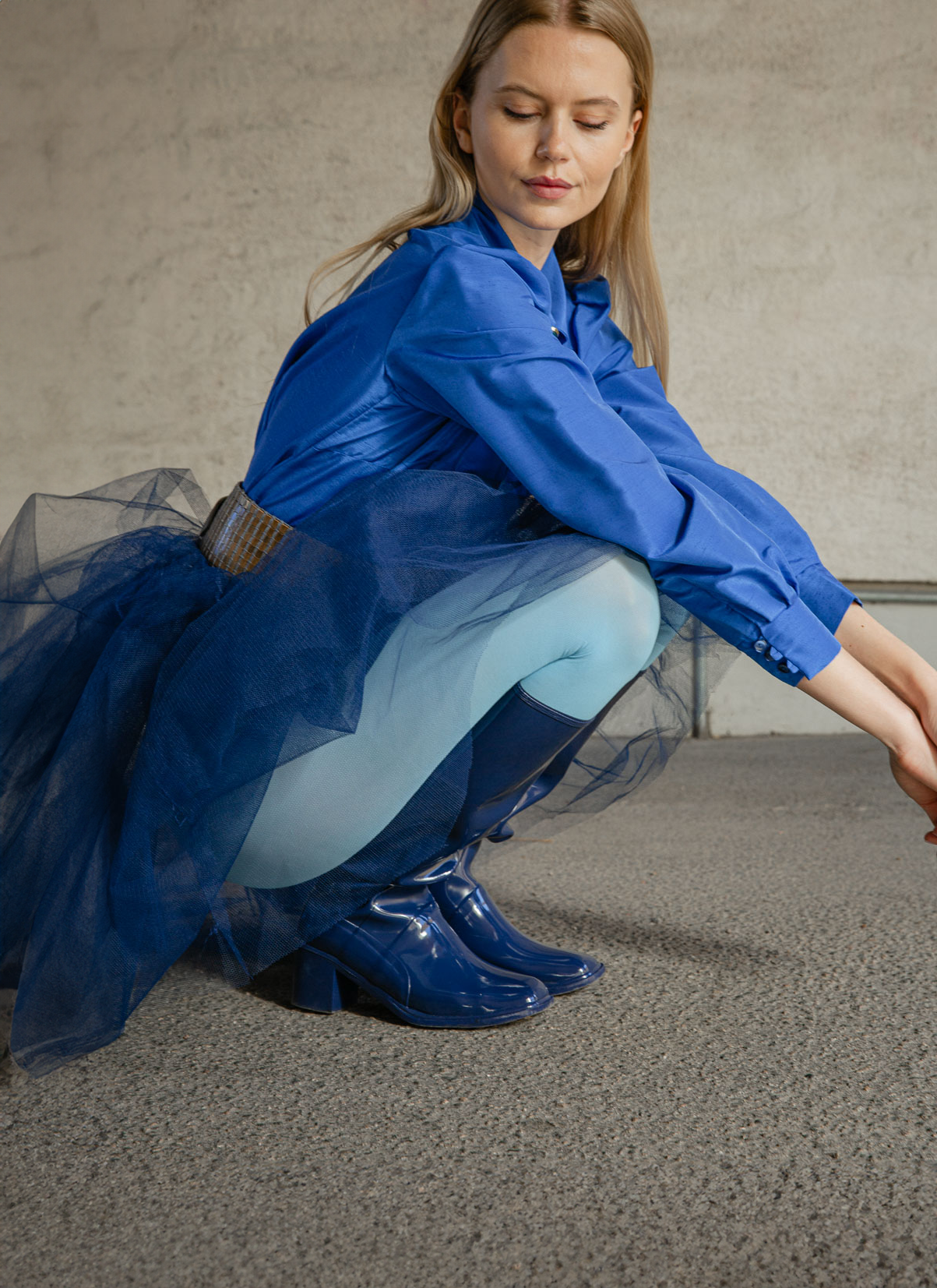 Model crouching in Karin Iris Blue knee-high rubber boots by ACQUO of Sweden, paired with a cobalt blue blouse, navy tulle skirt and light blue tights. Elegant waterproof boots with a sleek finish inspired by timeless Swedish design.
