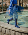 Model walking beside a fountain in Karin Iris Blue knee-high rubber boots by ACQUO of Sweden, styled with a flowing navy tulle skirt and light blue tights. The elegant blue waterproof boots feature a sleek reflective finish and timeless Swedish design.