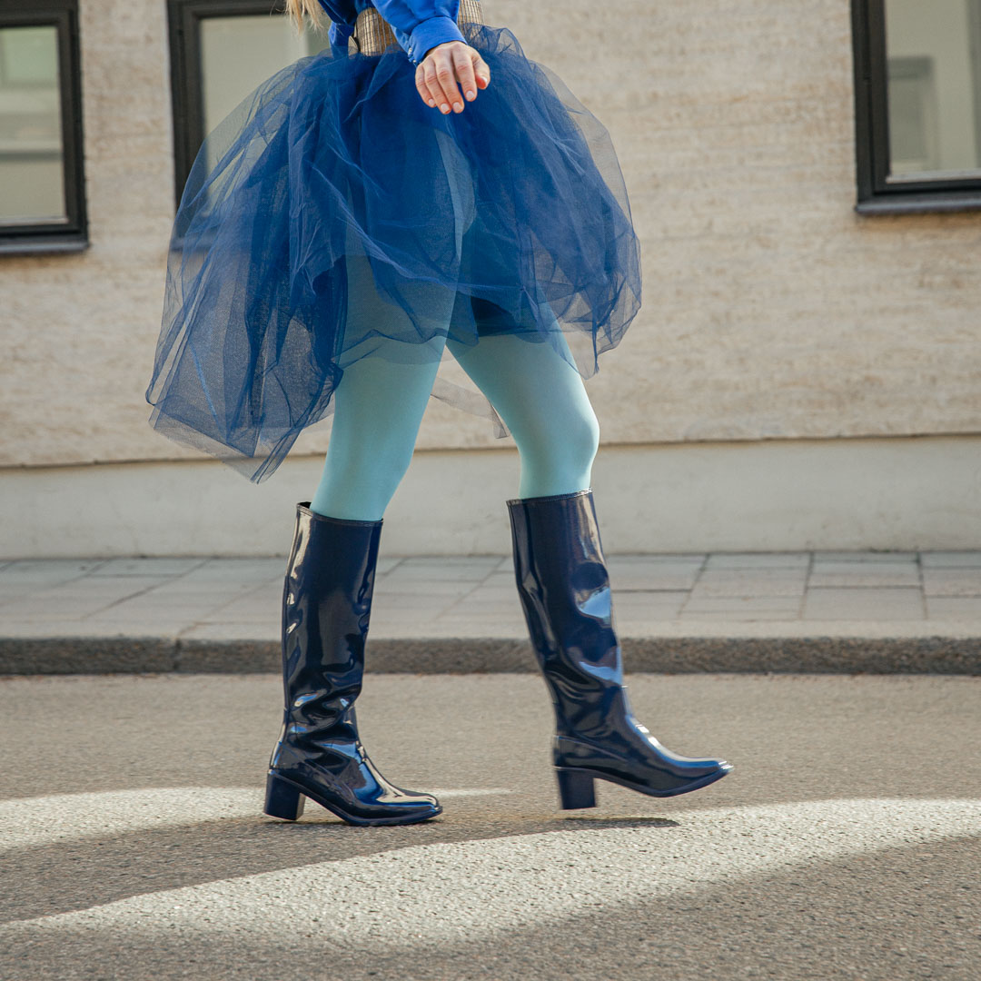 Model walking outdoors in Karin Iris Blue knee-high rubber boots by ACQUO of Sweden, styled with a flowing navy tulle skirt and light blue tights. The elegant blue waterproof boots feature a sleek reflective finish and modern Scandinavian fashion aesthetic.