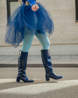Model walking outdoors in Karin Iris Blue knee-high rubber boots by ACQUO of Sweden, styled with a flowing navy tulle skirt and light blue tights. The elegant blue waterproof boots feature a sleek reflective finish and modern Scandinavian fashion aesthetic.
