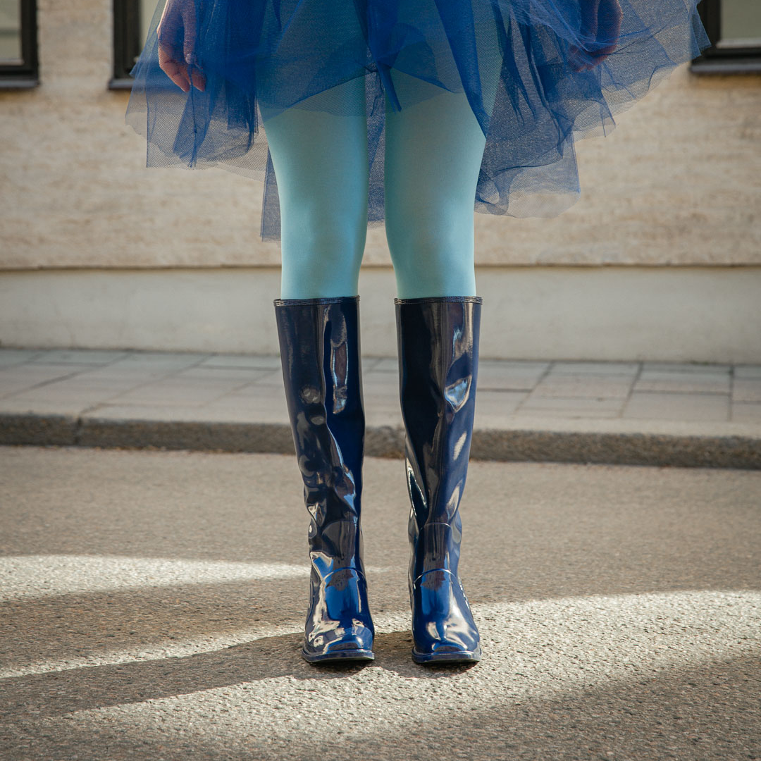 Front view of Karin Iris Blue knee-high rubber boots by ACQUO of Sweden styled with light blue tights and a navy tulle skirt outdoors. The elegant blue waterproof boots feature a sleek reflective finish and contemporary Scandinavian design.