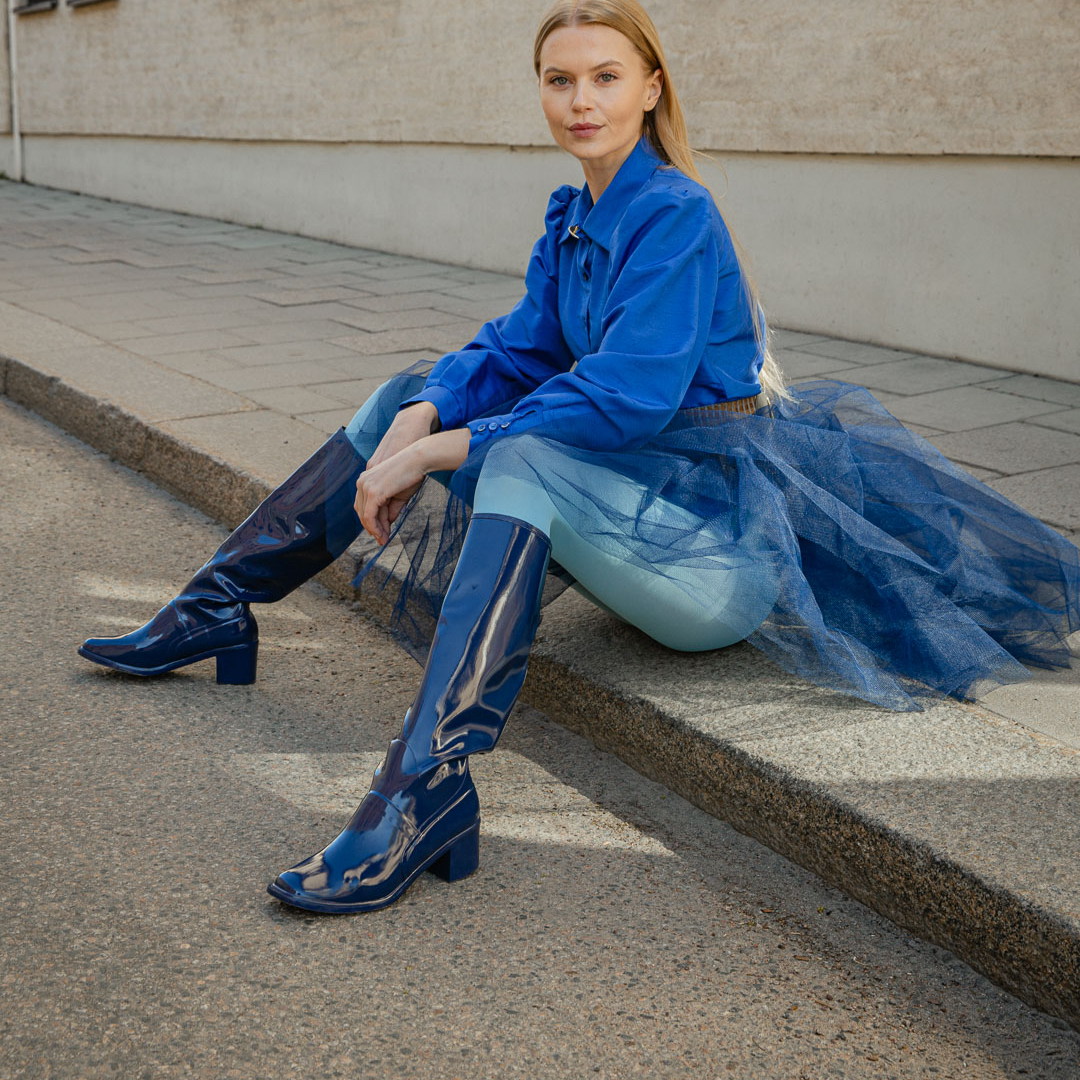 Model sitting outdoors in Karin Iris Blue knee-high rubber boots by ACQUO of Sweden, styled with a cobalt blue blouse, navy tulle skirt and light blue tights. The elegant blue waterproof boots feature a sleek reflective finish and timeless Swedish design