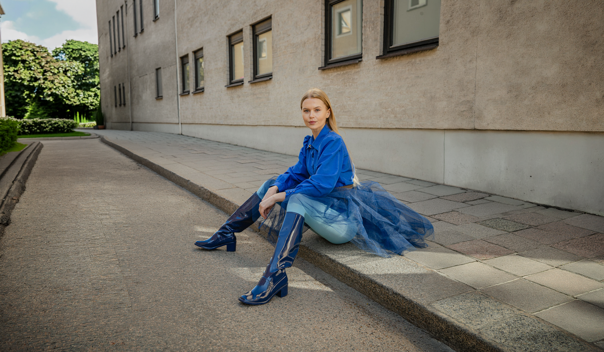 Model sitting on a city pavement wearing ACQUO of Sweden Karin Iris Blue knee-high natural rubber boots, styled with a blue jacket and layered tulle skirt in a Scandinavian urban setting