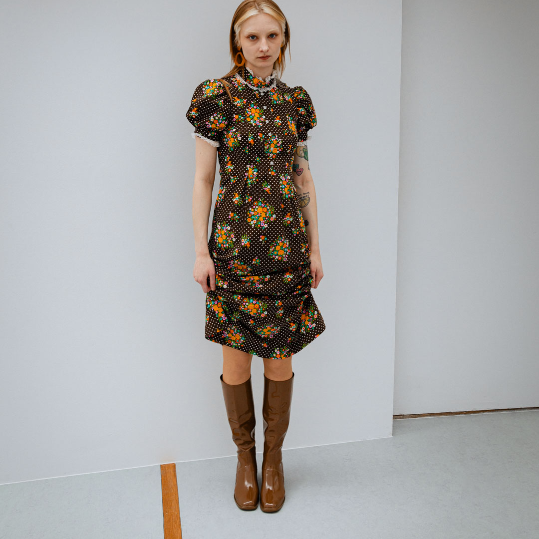 Woman wearing Karin Walnut Brown knee-high boots by ACQUO of Sweden paired with a vintage-inspired floral dress in a minimalist studio setting. Elegant waterproof boots with refined Swedish design, square toes and a timeless feminine silhouette.