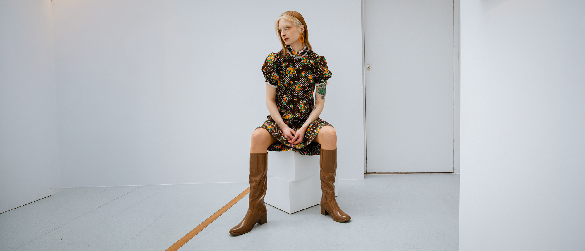Model sitting indoors wearing ACQUO of Sweden Karin Walnut Brown knee-high natural rubber boots, styled with a floral dress in a minimalist Scandinavian setting