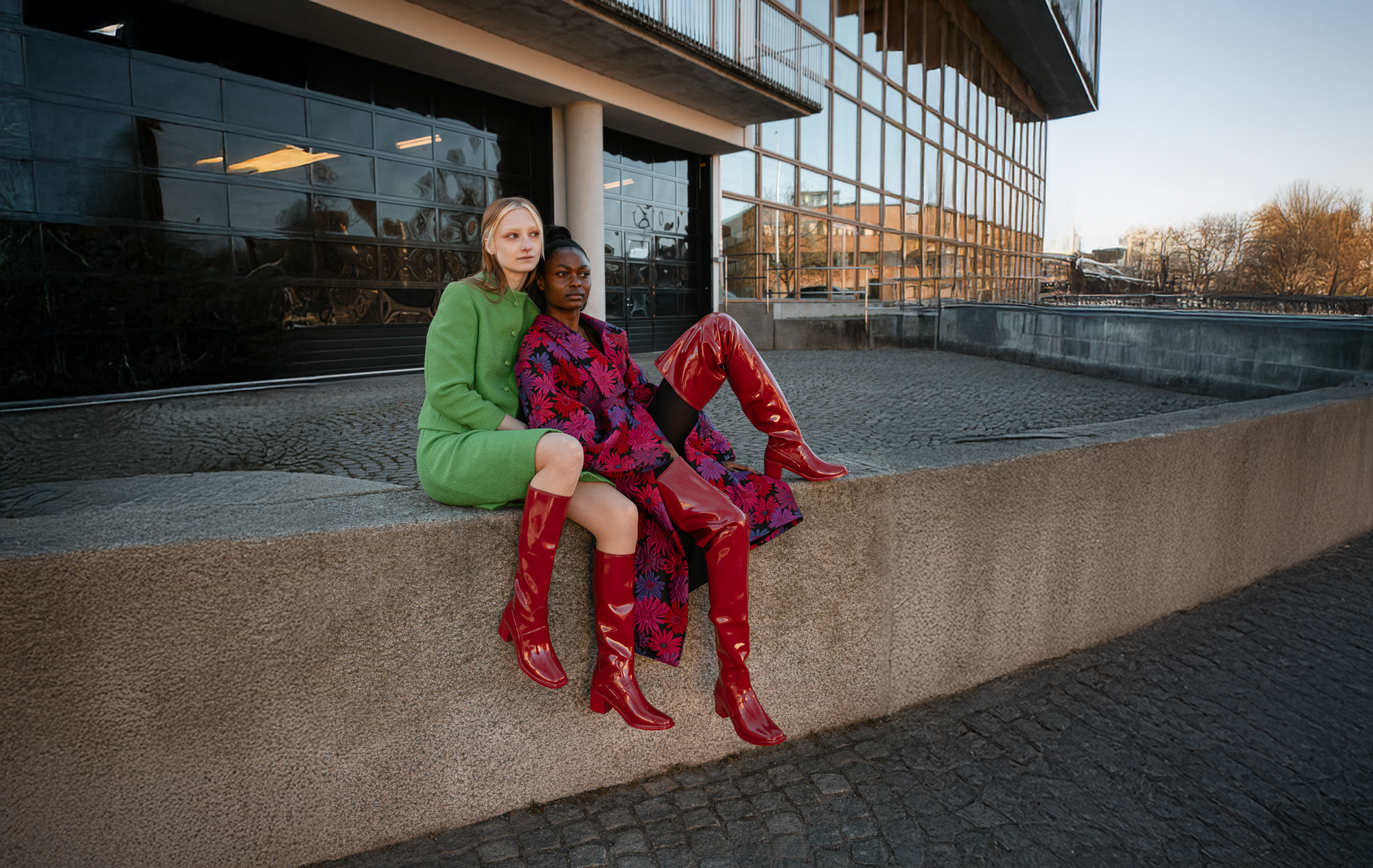 Two women wearing cherry red knee-high and over-the-knee boots by ACQUO of Sweden sitting in an urban architectural setting. Contemporary waterproof boots with square toes, block heels and minimalist Swedish design styled with bold fashion looks