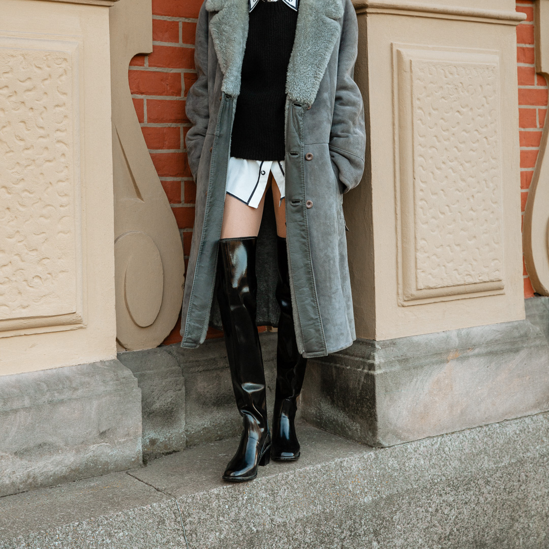 Model wearing ACQUO of Sweden over-the-knee glowing black boots, styled with a white shirt dress, black knit vest, and long grey shearling coat – a chic layered look combining modern fashion with classic Scandinavian outerwear