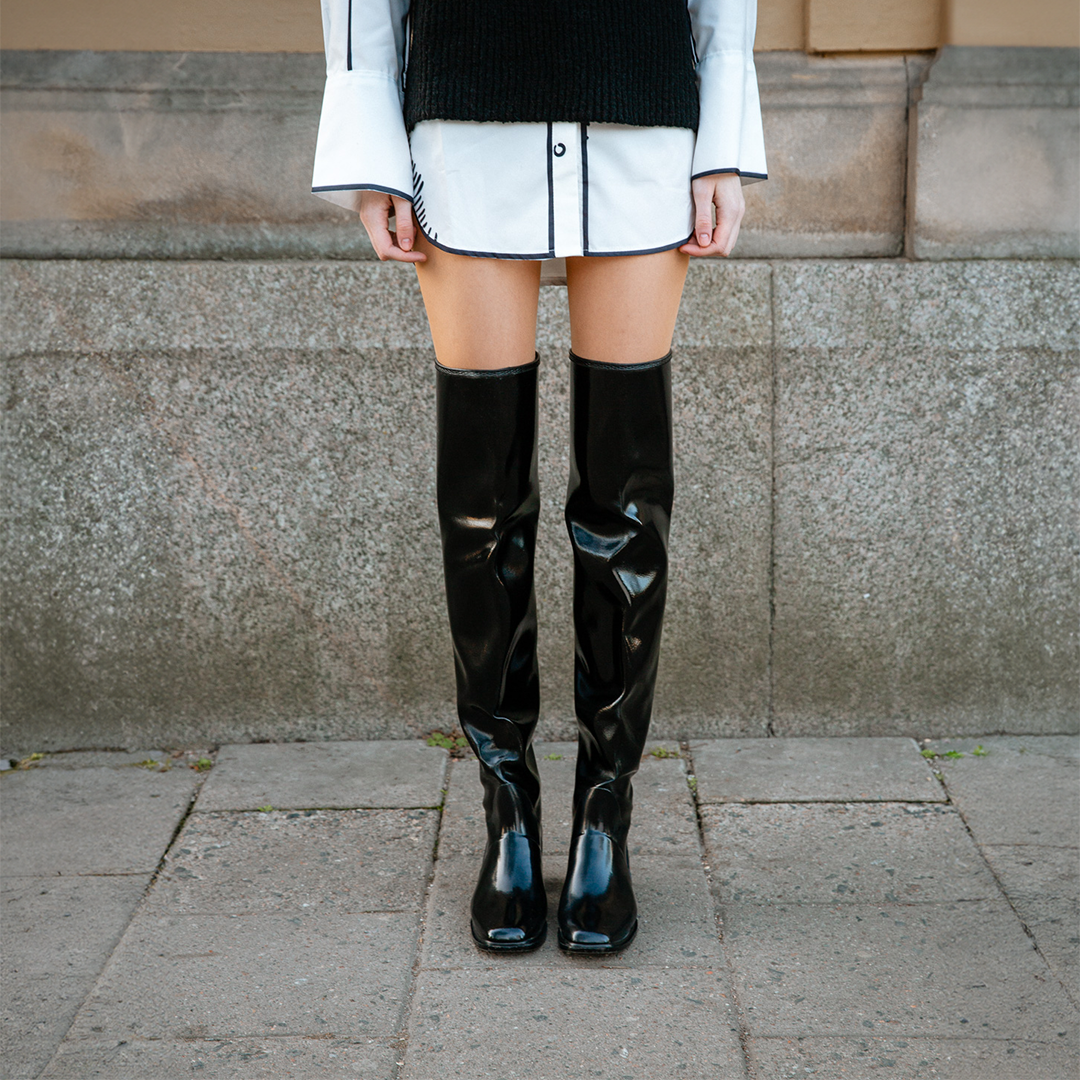 Close-up of model wearing ACQUO of Sweden over-the-knee glowing black boots, styled with a white shirt dress and black knitted vest – modern, statement footwear with a Scandinavian street style edge