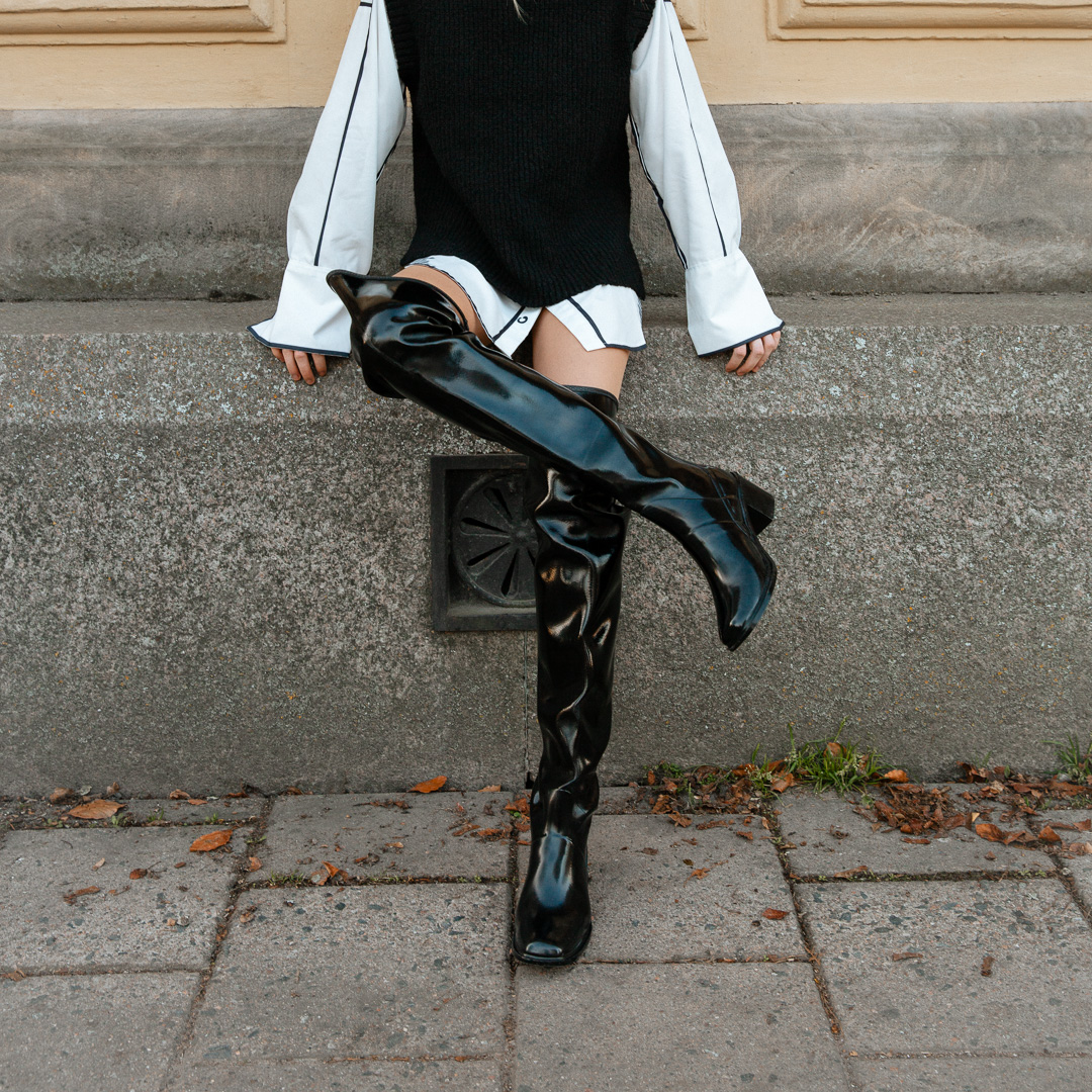 Model wearing ACQUO of Sweden over-the-knee glowing black boots, styled with a white oversized shirt, black knitted vest, and relaxed pose against a textured urban wall – a bold and modern Scandinavian fashion look