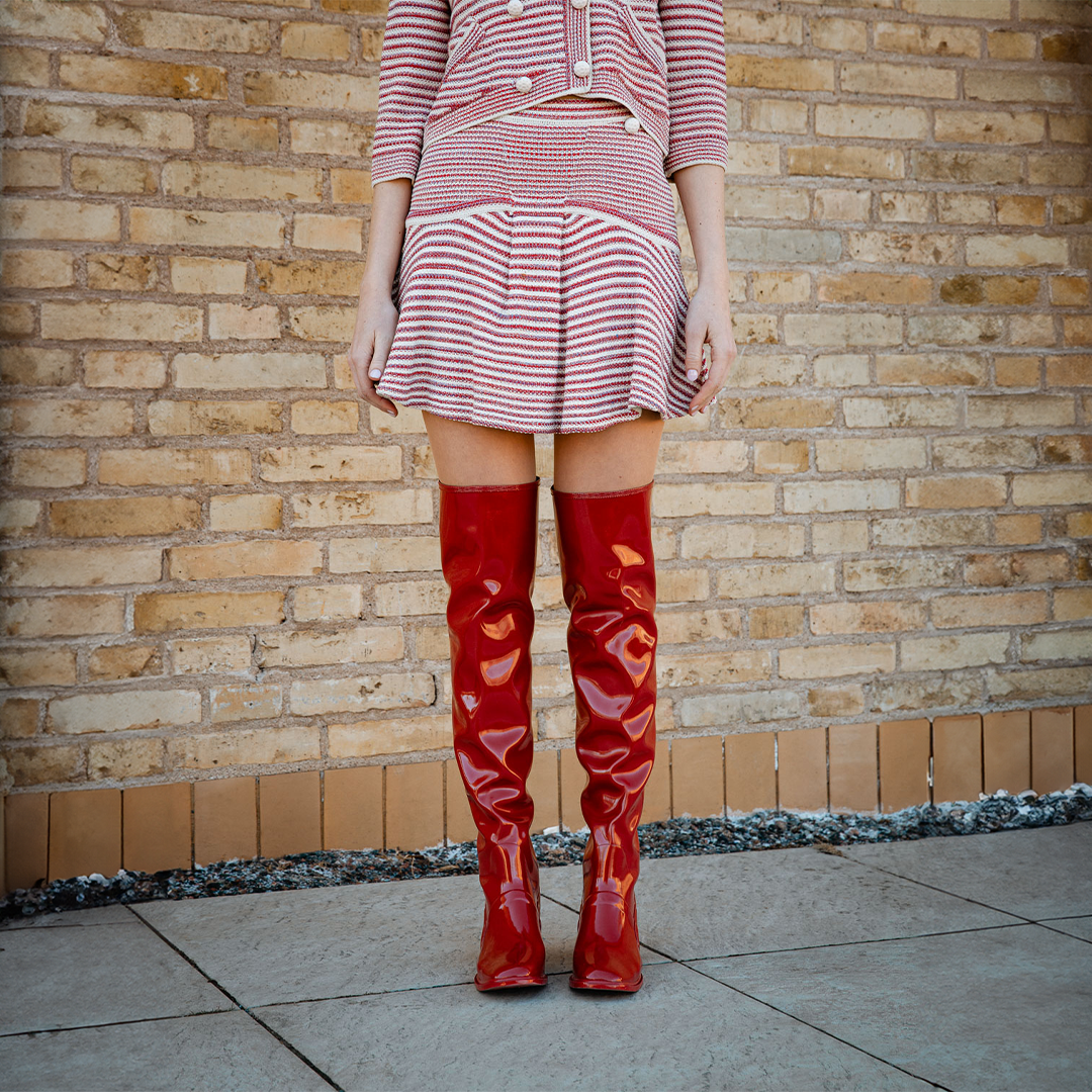 Model wearing Lisa Cherry Red over-the-knee boots by ACQUO of Sweden with a textured skirt suit against a brick wall. Elegant waterproof boots featuring bold Swedish design and contemporary Scandinavian fashion.