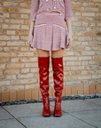 Model wearing Lisa Cherry Red over-the-knee boots by ACQUO of Sweden with a textured skirt suit against a brick wall. Elegant waterproof boots featuring bold Swedish design and contemporary Scandinavian fashion.
