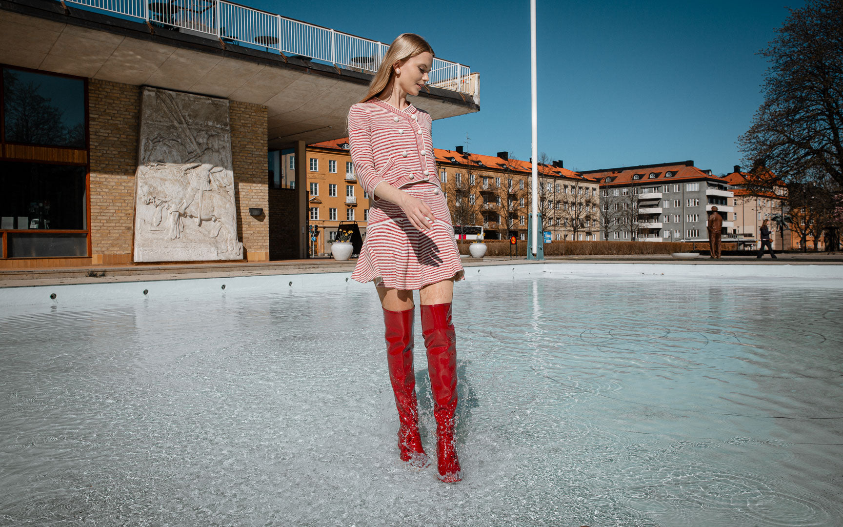 Model walking through shallow water in red knee-high natural rubber boots from ACQUO of Sweden, creating splashes with each step while wearing a red and white striped dress on a sunny day