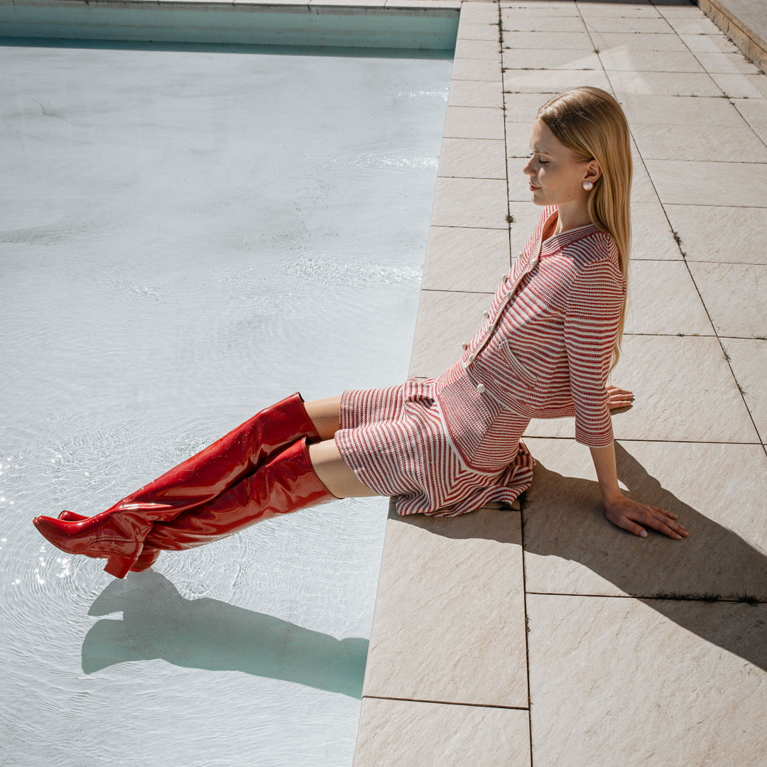 Model relaxing by a pool wearing Lisa Cherry Red over-the-knee boots by ACQUO of Sweden with a textured striped dress. Waterproof statement boots featuring elegant Swedish design and contemporary Scandinavian style.
