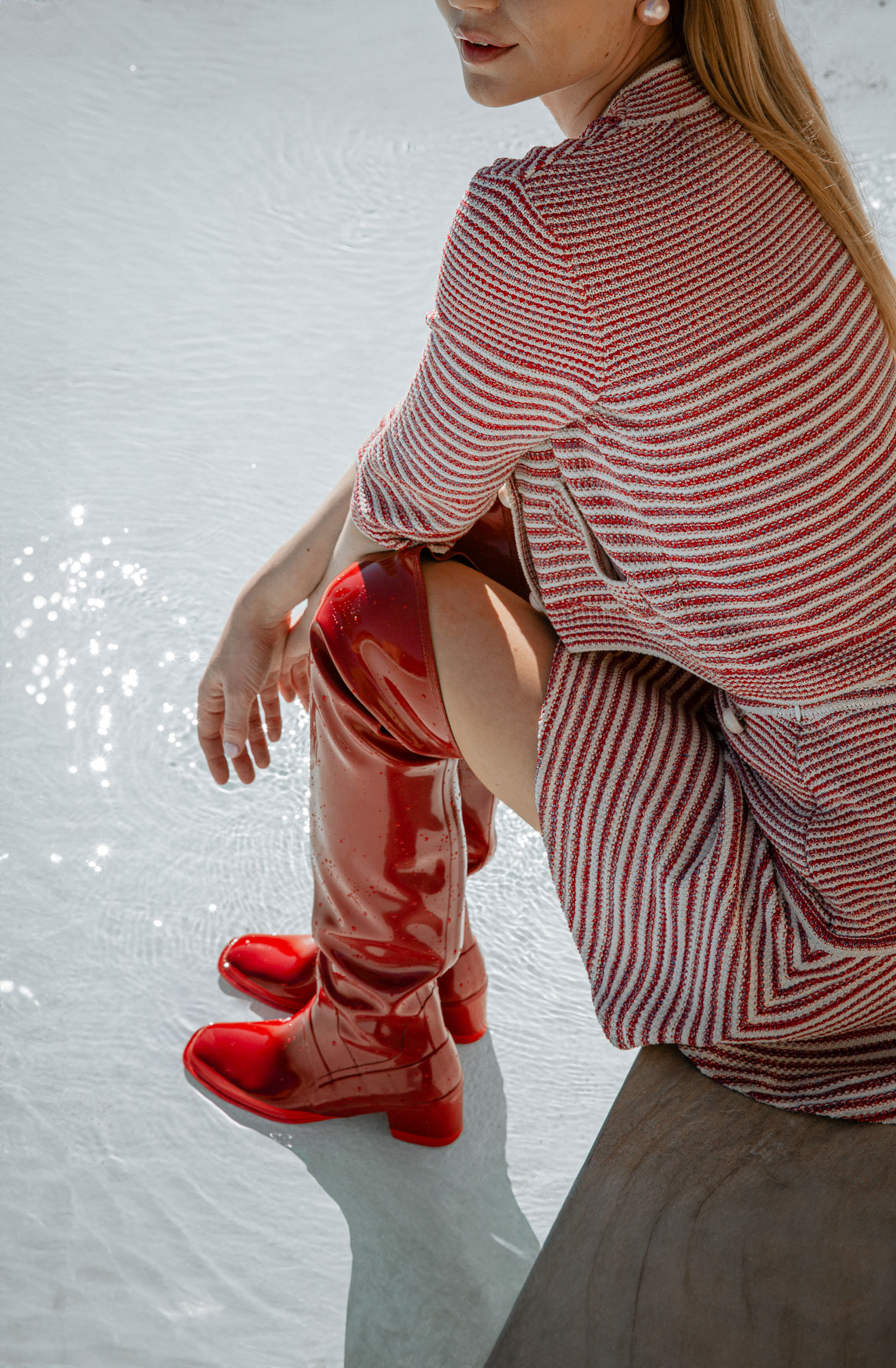 Model wearing Lisa Cherry Red knee-high boots by ACQUO of Sweden while sitting beside a pool in a striped knit outfit. Waterproof boots with elegant Swedish design and modern Scandinavian fashion styling.