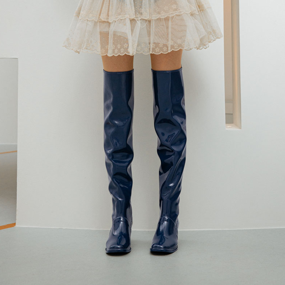 Front view of a model wearing Lisa Iris Blue boots by ACQUO of Sweden. The image highlights iris blue knee-high rubber boots with a sleek reflective finish and square toe silhouette, styled with a layered ivory tulle skirt in a minimalist indoor setting.
